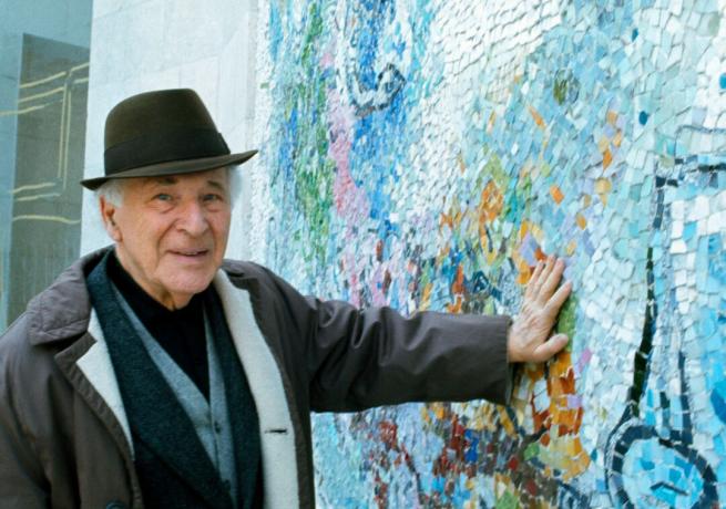 Kunstenaar Marc Chagall draagt ​​een hoed en drukt zijn hand tegen een muur met blauwe mozaïekontwerpen.