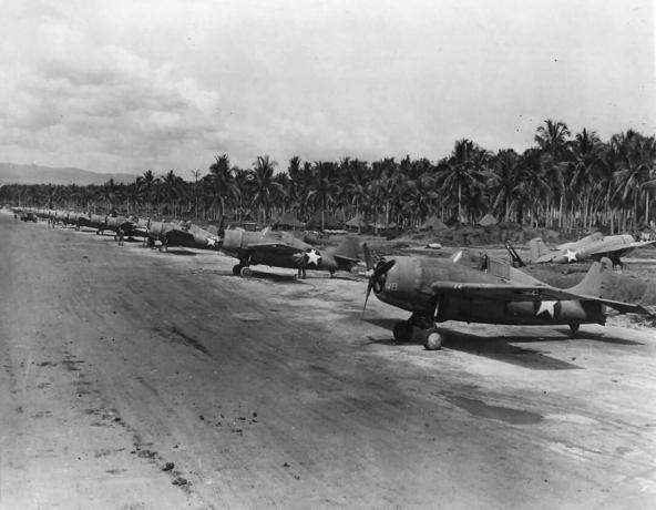 Rij van F4F Wildcats zitten langs een landingsbaan in een tropische omgeving.