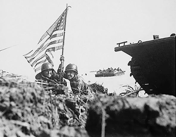 Twee soldaten met een Amerikaanse vlag op een strand naast een bijgehouden voertuig.